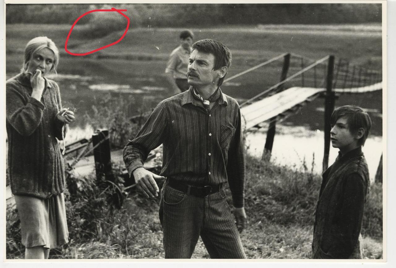 Black and white film still of people standing near a river and suspension bridge, with background trees highlighted.